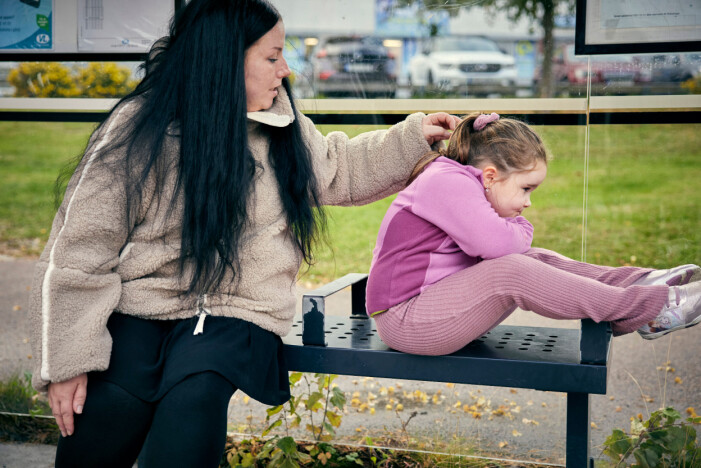 Även om det är bättre idag blir Alicia fortfarande lite rädd när en moped åker förbi, berättar mamma Sandra. Mamma Sandra klappar om lilla Alicia som sitter på en bänk i en busskur och ser orolig ut.