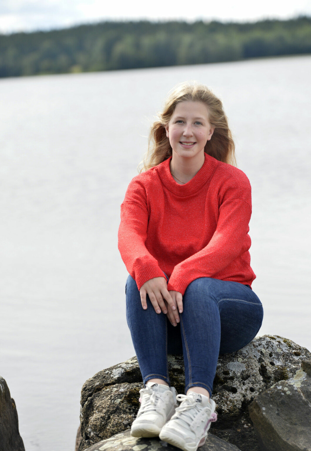 Filippa i röd tröja och blå jeans sitter på en sten vid en sjö
