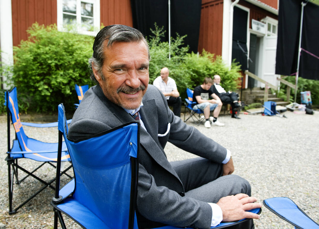 Kjell Bergqvist sitter på en campingstol framför ett rött hus