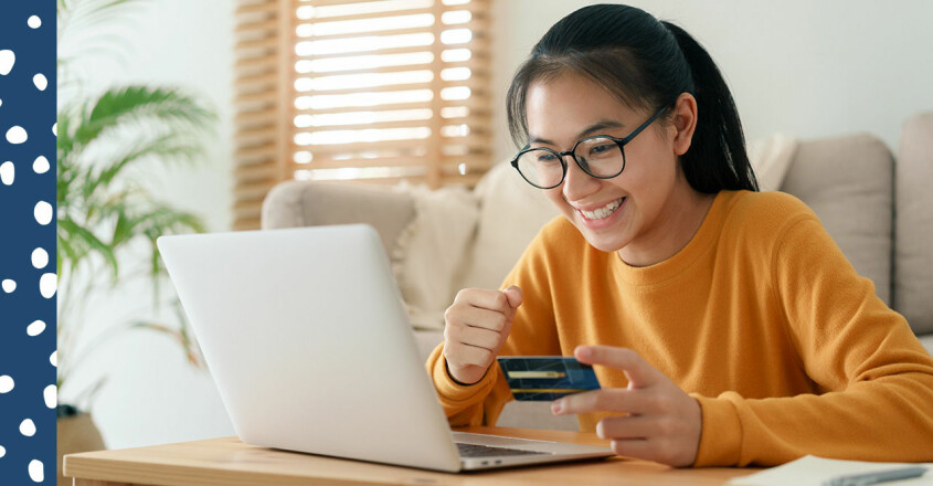 Ung kvinna håller i ett bankomatkort framför en dator och ser glad ut